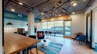 Located at 3883 S Moody Ave in Portland, Oregon, this single-floor amenity space blends industrial and contemporary design into a polished, functional environment. The open lounge area anchors the space with herringbone hardwood flooring, exposed concrete ceilings with structural columns, and a striking sculptural black pendant light fixture that serves as a dramatic focal point. Plush seating arrangements, including a deep navy wingback chair and a vibrant marbled area rug, create an inviting atmosphere for casual gatherings or focused work. Large floor-to-ceiling windows and a glass door fill the space with natural light and offer views of the surrounding Portland landscape. A distinctive word-search mural wall featuring hidden Portland-themed words adds a playful, locally inspired design element to the dining and lounge nook, which is furnished with upholstered booth seating and warm wood tables beneath matte black pendant lights. A dedicated conference room provides a more private setting, equipped with a long oak table, teal upholstered chairs, built-in display screen with wood surround, geometric linear pendant lighting, and a floor-to-ceiling window overlooking city views. A smaller meeting alcove, accessible via a sliding barn door, offers additional flexible workspace. Throughout, the design balances urban edge with warm, welcoming finishes.