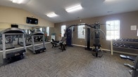 Located at 7113 Cape Harbor Dr in Wilmington, NC, this dedicated fitness center offers a well-equipped, single-floor workout facility designed to support a comprehensive exercise routine. The space features vaulted ceilings in select areas, adding an open and airy feel, while neutral-toned walls and patterned carpet flooring create a clean, functional environment throughout.

The cardio section is stocked with multiple treadmills, including a Landice model, as well as stationary and recumbent bikes, providing a variety of low-impact and high-intensity options. A multi-station cable machine and a dedicated strength-training apparatus anchor the free weights area, which also includes a dumbbell rack and a flat workout bench for versatile strength training.

Large wall-mounted mirrors line portions of the room, visually expanding the space and allowing users to monitor their form. Natural light filters in through several windows, complementing the overhead fluorescent lighting. A wall-mounted flat-screen television offers entertainment during workouts. Additional amenities include water coolers for hydration and a medicine ball rack for functional fitness exercises. A dedicated entryway with double doors provides easy access to the facility. The overall layout is thoughtfully arranged to maximize floor space and accommodate multiple users simultaneously.