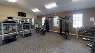Located at 7113 Cape Harbor Dr in Wilmington, NC, this dedicated fitness center offers a well-equipped, single-floor workout facility designed to support a comprehensive exercise routine. The space features vaulted ceilings in select areas, adding an open and airy feel, while neutral-toned walls and patterned carpet flooring create a clean, functional environment throughout.

The cardio section is stocked with multiple treadmills, including a Landice model, as well as stationary and recumbent bikes, providing a variety of low-impact and high-intensity options. A multi-station cable machine and a dedicated strength-training apparatus anchor the free weights area, which also includes a dumbbell rack and a flat workout bench for versatile strength training.

Large wall-mounted mirrors line portions of the room, visually expanding the space and allowing users to monitor their form. Natural light filters in through several windows, complementing the overhead fluorescent lighting. A wall-mounted flat-screen television offers entertainment during workouts. Additional amenities include water coolers for hydration and a medicine ball rack for functional fitness exercises. A dedicated entryway with double doors provides easy access to the facility. The overall layout is thoughtfully arranged to maximize floor space and accommodate multiple users simultaneously.