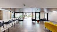 Located at 3170 W Olympic Blvd in Los Angeles, this single-floor communal space combines a fully equipped kitchen and a thoughtfully designed living room into one cohesive amenity area. Entry is made through sleek glass double doors, immediately signaling the modern aesthetic that defines the interior throughout.

The kitchen is a standout feature, showcasing light wood cabinetry that spans floor to ceiling, a generous white quartz island with bar seating for multiple guests, and stainless steel appliances including a double wall oven and a French door refrigerator. A wood-paneled ceiling adds warmth and depth, while large black-framed glass doors open directly to an outdoor courtyard, flooding the space with natural light and providing seamless indoor-outdoor flow.

The living room area is equally well-appointed, with large-format stone tile flooring transitioning to patterned carpeting in the lounge zone. A wall-mounted large-screen television serves as the focal point, complemented by a curated mix of seating including gray upholstered armchairs, a bold yellow sofa, and a rich walnut coffee table. A built-in fireplace with light wood surround cabinetry adds an inviting focal element, while recessed lighting and a wood-clad ceiling contribute to the warm, sophisticated ambiance throughout.