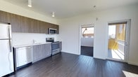 This single-floor space showcases a clean, modern design with an open and functional layout. The kitchen is a standout feature, equipped with stainless steel appliances including an electric range and over-the-range microwave, complemented by two-toned cabinetry — warm wood-finished upper cabinets paired with light gray lower cabinets. Light-colored countertops provide a bright contrast, while a single-basin sink with a contemporary faucet adds practicality. The crisp white walls throughout the space create a fresh, airy atmosphere, further enhanced by recessed and track lighting on the ceiling. Rich, dark hardwood flooring extends across the open area, adding warmth and a sense of continuity to the space. A doorway off the main area leads to an adjoining room, which features a window with an exterior view, allowing natural light to filter in and offering a glimpse of the surrounding environment. The overall design emphasizes simplicity and functionality, with a neutral color palette and modern finishes that give the space a contemporary, move-in-ready feel.