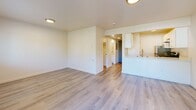 Located at 11020 SE Kent Kangley Rd in Kent, Washington, this single-floor unit offers a practical and well-appointed layout across a compact but functional footprint. The open-concept kitchen and living area serves as the heart of the home, featuring light wood-style flooring that extends throughout the space. The kitchen is equipped with white cabinetry, a stainless steel undermount sink with a contemporary gooseneck faucet, light quartz-style countertops, a stainless steel refrigerator, and a gas range with overhead ventilation — providing a clean, modern aesthetic. A peninsula counter separates the kitchen from the living area, adding both workspace and a visual boundary between the two zones. The living area is bright and airy, benefiting from a sliding glass door that opens to a private balcony overlooking lush, mature trees, complemented by an additional window that invites natural light. Baseboard heating runs along the perimeter walls. The full bathroom is finished in a clean, neutral palette with light wood-style flooring, a white vanity with storage, and a tub/shower combination. A dedicated closet provides storage, and the overall design maintains a consistent, updated look with white walls and modern hardware throughout.
