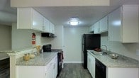 This single-story space centers around a well-appointed galley-style kitchen that maximizes efficiency through its classic parallel layout. The kitchen is outfitted with crisp white cabinetry on both upper and lower levels, providing ample storage throughout. Granite-look countertops run along both sides of the kitchen, offering generous prep space for cooking and meal preparation. Black appliances, including an electric range with overhead ventilation, a refrigerator, and a dishwasher, create a consistent and cohesive aesthetic against the white cabinetry. A stainless steel sink with a modern faucet is positioned along one counter run, adding a practical and clean touch. The kitchen features durable dark hardwood-style flooring that contrasts attractively with the lighter cabinetry and countertops. Overhead, a flush-mount ceiling light provides bright, even illumination throughout the space. The textured ceiling adds a subtle dimensional quality to the room. A door at the far end of the kitchen provides access to an adjacent area, contributing to the overall flow of the layout. The space is practical, clean, and thoughtfully organized for everyday use.