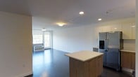 Located at 10 DeKalb Avenue in White Plains, New York, this single-floor residence offers a clean, contemporary design with an open-concept floor plan that seamlessly connects the kitchen and living areas. The kitchen is equipped with modern stainless steel appliances, including a full-size refrigerator with an in-door dispenser, and features sleek upper cabinetry in a light finish. A centrally positioned kitchen island with a warm wood base and white countertop serves as both a functional workspace and a natural gathering point. The flooring throughout is a rich, dark hardwood that adds depth and contrast to the bright, white walls and ceilings. Recessed lighting and flush-mount ceiling fixtures provide consistent illumination across the open living space. A large window with a roller shade allows for generous natural light and offers views of the surrounding greenery, while a glass door leads to an outdoor area, enhancing the sense of openness. The living area is spacious and versatile, providing ample room for various furniture arrangements. Clean architectural lines and a neutral color palette give the space a modern, move-in-ready aesthetic suited for comfortable everyday living in the heart of White Plains.