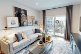 This 2-bedroom, 2-bathroom residence is designed with a modern aesthetic and a functional open-concept floor plan. The kitchen is a standout feature, outfitted with gray shaker-style cabinetry, white quartz countertops, a central island with bar seating, stainless steel appliances including a gas range, built-in microwave, and French door refrigerator, all set atop warm-toned hardwood flooring. The kitchen flows seamlessly into a dedicated dining area and the living room, creating an ideal space for entertaining. The living room is bright and airy, anchored by large sliding glass doors that open onto a private balcony with outdoor views. The spacious primary bedroom features warm, earthy wall tones, recessed lighting, hardwood flooring, and a large window that fills the room with natural light. It connects directly to a well-appointed en-suite bathroom featuring a walk-in glass shower, tile flooring, a wood-toned vanity, and wall-mounted storage. The second bedroom is equally comfortable, and the second bathroom mirrors the contemporary design with a wood-finish vanity, tile flooring, and clean white surfaces. The overall ambiance throughout the residence is modern, clean, and inviting, with consistent finishes tying each space together cohesively.