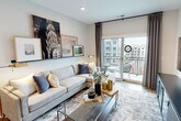 This 2-bedroom, 2-bathroom residence is designed with a modern aesthetic and a functional open-concept floor plan. The kitchen is a standout feature, outfitted with gray shaker-style cabinetry, white quartz countertops, a central island with bar seating, stainless steel appliances including a gas range, built-in microwave, and French door refrigerator, all set atop warm-toned hardwood flooring. The kitchen flows seamlessly into a dedicated dining area and the living room, creating an ideal space for entertaining. The living room is bright and airy, anchored by large sliding glass doors that open onto a private balcony with outdoor views. The spacious primary bedroom features warm, earthy wall tones, recessed lighting, hardwood flooring, and a large window that fills the room with natural light. It connects directly to a well-appointed en-suite bathroom featuring a walk-in glass shower, tile flooring, a wood-toned vanity, and wall-mounted storage. The second bedroom is equally comfortable, and the second bathroom mirrors the contemporary design with a wood-finish vanity, tile flooring, and clean white surfaces. The overall ambiance throughout the residence is modern, clean, and inviting, with consistent finishes tying each space together cohesively.