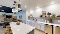 This thoughtfully designed single-floor amenity space offers a generous open-plan layout that seamlessly integrates multiple functional zones for socializing and recreation. The kitchenette serves as a central hub, featuring crisp white shaker cabinets, white quartz countertops, a stainless steel sink with a matte black faucet, and a geometric tile backsplash in soft grey. Open wood shelving adds warmth and display space, while two under-counter refrigerators provide convenient beverage storage. A boldly colored teal kitchen island with bar seating anchors the space and creates a natural divide between the kitchen and dining areas. An eclectic cluster of pendant lights in mixed black, white, and gold tones illuminates the island and adds visual interest overhead. The dining area accommodates a large communal wood table surrounded by black spindle chairs, positioned beneath a striking dark-painted ceiling section that adds depth and contrast. The lounge area features a comfortable sectional sofa, rounded leather accent chairs, and a large area rug, all oriented toward a wall-mounted flat-screen TV framed by open industrial-style shelving. A billiard table is visible in the background, rounding out the entertainment offerings. Light wood-look flooring flows throughout, unifying the space with a clean, contemporary aesthetic.