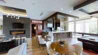 Located at 1000 Antietam Creek Dr in Fayetteville, NC, this single-floor community clubhouse offers a thoughtfully designed gathering space with a range of amenities. The open-concept layout seamlessly connects multiple functional zones, creating an inviting atmosphere for socializing and entertainment. A sleek bar counter with stylish bar stools anchors the space, complemented by shelving and appliances that make it well-suited for hosting and casual gatherings. Round dining tables paired with modern white chairs are arranged throughout the lounge area, providing comfortable seating options for residents. A contemporary fireplace set into a dark stone accent wall adds warmth and visual interest to one side of the room. The space is illuminated by a combination of recessed ceiling lights and abundant natural light streaming through large windows. A dedicated theater room, clearly marked with illuminated signage, offers a private cinematic experience just steps away from the main lounge. The interior design blends warm wood flooring with contrasting dark ceiling panels and a rich terracotta accent wall, creating a modern yet welcoming ambiance. The overall layout encourages both intimate conversation and larger community gatherings.