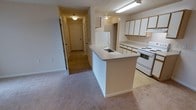 This single-floor residence features a functional, open-concept layout that connects the kitchen directly to the main living area. The kitchen is equipped with white appliances, including a freestanding electric range and a full-sized refrigerator, complemented by light wood-trimmed upper cabinetry offering ample storage. A peninsula with a built-in sink serves as a natural dividing point between the kitchen and the adjacent carpeted living space, providing both additional prep space and a casual bar-style area.

The flooring transitions from resilient vinyl or linoleum in the kitchen to neutral-toned wall-to-wall carpeting throughout the living area and hallway, contributing to a warm and cohesive feel. The hallway extending from the main space leads to multiple doors, suggesting several rooms such as bedrooms and bathrooms, offering a well-organized, private layout. Overhead fluorescent and flush-mount ceiling fixtures provide consistent artificial lighting throughout. Neutral beige and white tones dominate the interior palette, creating a clean, understated aesthetic. White baseboards and trim provide a crisp finish throughout the space.