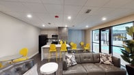 This single-floor space offers an open-plan layout that seamlessly integrates a kitchen, dining area, and lounge into one cohesive environment. The kitchen is outfitted with stainless steel appliances, including a microwave and range, alongside a black refrigerator and upper cabinetry in a light neutral finish. A stone-tiled accent wall serves as a striking backsplash, adding texture and visual depth. A white breakfast bar extends from the kitchen, accompanied by vibrant yellow bar stools that introduce a bold pop of color throughout the space. The dining area continues this energetic palette with matching yellow chairs arranged around sleek tables. The lounge area features a contemporary grey leather sofa accented with patterned throw pillows, positioned beside a small round white side table. Dark hardwood-style flooring runs throughout, grounding the bright accents with a warm contrast. Recessed lighting and a drop tile ceiling ensure even illumination across the space. A large sliding glass door or window panel along one wall floods the interior with natural light and provides a clear view of an outdoor pool area, enhancing the overall open and inviting ambiance.