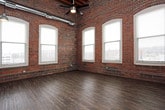 This loft-style unit masterfully combines industrial heritage with contemporary living. The open-plan living and kitchen area is defined by dramatic soaring ceilings with exposed wooden beams and ductwork overhead, creating a striking visual depth. Rich, dark hardwood-style plank flooring runs throughout the main living space, complemented by original exposed brick walls that lend warmth and historic character. Arched windows along the brick walls flood the space with natural light and offer views of the surrounding urban landscape.

The kitchen is outfitted with rich espresso-toned cabinetry, light countertops, black appliances including a refrigerator, range, and dishwasher, and is accented by the same exposed brick wall alongside a window. Pendant lighting adds an industrial-modern touch above the workspace.

The bedroom continues the loft aesthetic with soaring ceilings, exposed brick, arched windows, plush carpeting, and a ceiling fan. A closet provides convenient storage, and the room connects directly to the bathroom.

The bathrooms feature tile flooring, a combination tub and shower with chrome fixtures and a curtain rod, a dark vanity with a light countertop, and clean, neutral wall tones. The overall ambiance throughout is urban, open, and full of character.