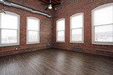 This loft-style unit masterfully combines industrial heritage with contemporary living. The open-plan living and kitchen area is defined by dramatic soaring ceilings with exposed wooden beams and ductwork overhead, creating a striking visual depth. Rich, dark hardwood-style plank flooring runs throughout the main living space, complemented by original exposed brick walls that lend warmth and historic character. Arched windows along the brick walls flood the space with natural light and offer views of the surrounding urban landscape.

The kitchen is outfitted with rich espresso-toned cabinetry, light countertops, black appliances including a refrigerator, range, and dishwasher, and is accented by the same exposed brick wall alongside a window. Pendant lighting adds an industrial-modern touch above the workspace.

The bedroom continues the loft aesthetic with soaring ceilings, exposed brick, arched windows, plush carpeting, and a ceiling fan. A closet provides convenient storage, and the room connects directly to the bathroom.

The bathrooms feature tile flooring, a combination tub and shower with chrome fixtures and a curtain rod, a dark vanity with a light countertop, and clean, neutral wall tones. The overall ambiance throughout is urban, open, and full of character.
