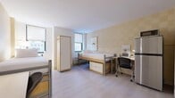 Located at 525 Lexington Avenue in the heart of Midtown Manhattan, New York, this double-occupancy room offers a practical and efficiently designed living space ideal for two residents. The main room features light wood-look flooring and warm, neutral-toned walls that create a bright and welcoming atmosphere. Two loft-style beds are positioned to maximize floor space, each elevated to allow for built-in drawer storage underneath — a smart solution for keeping belongings organized in a compact setting. Each resident is provided with a dedicated study desk and ergonomic chair, with built-in lighting to support productivity. A full-height wardrobe with coat hooks offers additional storage and organization. Multiple windows allow natural light to fill the room, offering views of the surrounding Midtown streetscape. A colorful art print near the entryway adds a touch of personality to the space. The private bathroom is finished with classic cream-toned tile work from floor to ceiling, and includes a full soaking bathtub with shower, a granite-topped vanity, chrome fixtures, and safety grab bars. The entryway area provides additional functional space, complete with a compact refrigerator and easy access to the main entry door.