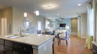 This two-story home offers a thoughtfully designed open-concept main level that seamlessly connects the kitchen, dining, and living areas into one cohesive, flowing space. The kitchen is appointed with granite countertops, dark cabinetry, and a central island with pendant lighting above, providing both functional workspace and a natural gathering point. Track lighting and recessed fixtures illuminate the area efficiently throughout.

The dining area sits just off the kitchen, accommodating a full dining table and chairs beneath a direct sightline to the living room beyond. Hardwood flooring runs continuously across the main level, lending warmth and visual continuity to the open layout.

The living room features a ceiling fan, a wall-mounted television, and large windows dressed with curtains that allow natural light to fill the space. A soft teal accent wall adds a subtle pop of color, complementing the neutral tones found throughout the home. A staircase is visible at the rear of the living area, leading to the second floor. The overall ambiance is bright, modern, and inviting, with a clean, coordinated aesthetic throughout.