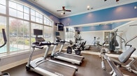 Located at 2305 Global Forum Blvd in Atlanta, GA, this single-floor fitness center offers a comprehensive workout environment designed for both cardio and strength training. The space is finished with soft blue walls accented by crisp white crown molding and baseboards, creating a bright, energizing atmosphere throughout. Large arched and grid-pane windows line the perimeter, flooding the interior with abundant natural light and providing views of the surrounding outdoor area, including a glimpse of a pool. Full-length mirrored walls on one side visually expand the space and allow users to monitor their form during exercise. The flooring consists of durable, dark rubberized material suited for high-traffic fitness use. Ceiling fans are installed throughout to promote airflow and comfort. The cardio section is well-stocked with multiple Spirit-brand treadmills and elliptical trainers, each equipped with individual mounted flat-screen monitors. The strength training area includes a multi-function cable machine, weight benches, and free weights. A set of elegant white French doors with glass panes connects separate sections of the facility, adding a refined architectural touch. Recessed and ceiling-mounted lighting supplements natural light for consistent illumination across all workout areas.