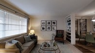 Located at 9400 Abercorn St in Savannah, Georgia, this single-floor residence offers a comfortable and well-appointed living environment with a cohesive, understated aesthetic. The living room serves as the heart of the space, showcasing soft, neutral walls that create a warm and inviting atmosphere. Plush carpeting underfoot adds to the sense of comfort, while a large window fitted with horizontal blinds and patterned drape panels allows for natural light to filter into the room. A built-in bookcase along one wall provides both functional storage and a display area, adding architectural character to the space. The room is thoughtfully arranged with a cushioned sofa, a classic leather accent chair, and a glass-topped coffee table, all anchored by a layered area rug. A curated gallery wall featuring six framed art prints lends a polished, finished look to the interior. The overall ambiance is calm and livable, reflecting a design sensibility that balances practicality with tasteful style. The single-story layout offers ease of accessibility and a cohesive flow throughout the home, making it a functional and comfortable place to call home in the heart of Savannah.