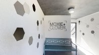 This single-floor space makes a strong first impression through its thoughtfully designed entrance area. The most striking feature is the white accent wall adorned with a pattern of hexagonal shapes in varying shades of gray, black, and white, creating a modern, geometric visual display. The "Moxie + Clover" logo is prominently displayed on the adjacent white wall, establishing a clear brand presence. A painted teal wooden bench with cross-back detailing sits beneath the signage, adding a touch of character and color to the otherwise neutral palette. The ceiling features an exposed concrete finish, lending an industrial yet polished aesthetic that contrasts effectively with the bright white walls. A flush-mount ceiling light provides ambient illumination, while a window near the bench allows natural light to filter into the space. The overall ambiance is clean, modern, and inviting, blending industrial and boutique design elements seamlessly. The space conveys a curated, brand-conscious atmosphere suited to a retail or studio environment.