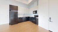This modern single-floor unit presents a clean, contemporary design throughout, unified by warm light wood flooring and crisp white walls. The open-concept living area is generously proportioned, offering a bright and airy atmosphere enhanced by a large multi-pane window that frames scenic outdoor views of the surrounding cityscape.

The kitchen is thoughtfully designed in an L-shaped layout, featuring a striking two-tone aesthetic with dark lower cabinetry and white upper cabinets set against a white tile backsplash. It is fully equipped with stainless and black appliance finishes, including a refrigerator, electric range, over-the-range microwave, and dishwasher, along with a single-basin sink. The kitchen flows naturally into the main living space, making it ideal for everyday functionality.

The bedroom is a calm, well-lit retreat with high ceilings, recessed lighting, and a window that provides natural light and urban views. A dedicated in-unit laundry closet houses a stacked washer and dryer, adding practical convenience.

The bathroom is finished with classic white subway-style tile surrounding a full soaking tub and shower combination, complemented by a modern vanity with an undermount sink, a large mirror, and wood-tone flooring that carries throughout the space.