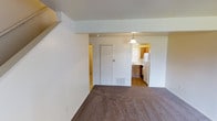 This two-story space offers a functional and straightforward layout designed for comfortable everyday living. The main level features a generously sized living area finished with wall-to-wall carpet in a neutral tone, providing a warm and understated foundation for the room. Clean white walls and a simple, open layout give the space a bright and airy feel.

A bifold closet door along one wall offers convenient built-in storage within the living area. A floor vent is positioned nearby, indicating an integrated heating and cooling system throughout the space. The living area flows naturally into the kitchen through an open passageway, maintaining a connected yet distinct separation between the two spaces.

The kitchen is appointed with warm wood-toned cabinetry, a white refrigerator, and a pendant light fixture that hangs above, adding a subtle decorative touch. A window in the kitchen allows natural light to filter into the space. Hard surface flooring in the kitchen contrasts with the carpeted living area, providing a practical transition between zones.

A hallway visible from the living area leads toward additional rooms and provides access to the staircase connecting the two floors, suggesting further functional space above.