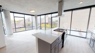 This contemporary urban residence offers a striking blend of industrial and modern design across a single level. Exposed concrete ceilings throughout lend an industrial character, while light-toned hardwood flooring and crisp white walls create a bright, refined atmosphere. Floor-to-ceiling windows wrap the living and sleeping areas, flooding the space with natural light and framing expansive views of the surrounding cityscape, including tree-lined streets and neighboring brick buildings.

The open-concept kitchen is a standout feature, equipped with gray shaker-style cabinetry, quartz countertops, a geometric tile backsplash, and a full suite of stainless steel appliances. A generously sized center island with a gas cooktop and a suspended stainless steel range hood serves as the kitchen's focal point, making it ideal for cooking and entertaining.

The bedrooms are spacious and airy, each benefiting from corner or wide-panel window configurations that maximize light and views. One bedroom includes a built-in closet with shelving. The two bathrooms feature gray vanities with quartz counters and large-format tile flooring; one includes a walk-in shower. A dedicated laundry closet with a stacked washer and dryer adds everyday convenience to this well-appointed urban home.