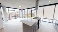 This contemporary urban residence offers a striking blend of industrial and modern design across a single level. Exposed concrete ceilings throughout lend an industrial character, while light-toned hardwood flooring and crisp white walls create a bright, refined atmosphere. Floor-to-ceiling windows wrap the living and sleeping areas, flooding the space with natural light and framing expansive views of the surrounding cityscape, including tree-lined streets and neighboring brick buildings.

The open-concept kitchen is a standout feature, equipped with gray shaker-style cabinetry, quartz countertops, a geometric tile backsplash, and a full suite of stainless steel appliances. A generously sized center island with a gas cooktop and a suspended stainless steel range hood serves as the kitchen's focal point, making it ideal for cooking and entertaining.

The bedrooms are spacious and airy, each benefiting from corner or wide-panel window configurations that maximize light and views. One bedroom includes a built-in closet with shelving. The two bathrooms feature gray vanities with quartz counters and large-format tile flooring; one includes a walk-in shower. A dedicated laundry closet with a stacked washer and dryer adds everyday convenience to this well-appointed urban home.