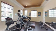 This fitness center offers a comprehensive workout environment designed to support a wide range of exercise routines. The space is anchored by a striking tray ceiling with a warm, contrasting accent color and recessed lighting that creates an inviting ambiance throughout. Crown molding adds a refined architectural touch, elevating the overall aesthetic beyond a typical gym setting.

Natural light flows in generously through multiple windows fitted with horizontal blinds, providing views of the surrounding greenery and keeping the space feeling open and energizing. Dark, durable rubber flooring covers the entire area, offering both safety and comfort underfoot during workouts.

The cardio section is well-stocked with Star Trac treadmills and elliptical trainers, complemented by upright and recumbent stationary bikes. A flat-screen television mounted on the wall adds entertainment options during cardio sessions. For strength training, the facility includes a multi-station cable machine, weight benches, free weights, and dedicated resistance equipment. A large wall mirror in the strength area enhances the sense of space while allowing users to monitor their form.

Motivational fitness posters adorn the walls, contributing to an encouraging workout atmosphere. A dedicated entry door provides convenient, direct access to the facility, making this fitness center a practical and polished amenity.