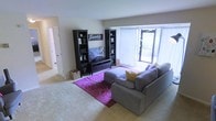 This single-floor space centers around a comfortable and thoughtfully arranged living room. The room is anchored by a large gray sectional sofa paired with a rectangular ottoman, both positioned atop a vibrant magenta floral-patterned area rug that adds a bold pop of color to the neutral carpeted flooring. A dark-finished entertainment center serves as the focal point of the main wall, flanked by two tall matching black bookshelves displaying decorative items and small potted plants. A flat-screen television is mounted within the entertainment unit, accompanied by a "Family" wall sign that adds a personal touch to the space. Natural light flows generously into the room through a wide window dressed with sheer white vertical blinds, offering a glimpse of outdoor greenery. The walls are finished in a soft, neutral tone that complements the overall warm and inviting atmosphere. A hallway visible to the left suggests additional rooms beyond the living area, with carpeted flooring continuing throughout. The space strikes a balance between functional layout and personalized décor, making it feel both livable and welcoming.
