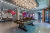 Located at 951 Glenwood Ave SE in Atlanta, GA, this single-floor space delivers a striking industrial-chic aesthetic that seamlessly blends raw architectural character with vibrant, artistic energy. Polished concrete floors stretch throughout, reflecting light and lending the interior an open, loft-like quality. Exposed ductwork and piping trace the ceiling above, reinforcing the industrial sensibility while adding visual depth overhead.

The space is anchored by a central game area showcasing a full-size pool table beneath a dramatic grid of Edison-style bulbs, creating a warm, focused glow. Surrounding exposed brick walls and large windows flood the area with natural light, connecting the interior to the outdoors. Cue racks and a chalkboard panel nearby complete the game room setup.

Adjacent to the game area, a lounge zone features a deep, cushioned sectional sofa paired with a wall-mounted television, all set against a sweeping, large-scale abstract mural rendered in bold pinks, reds, and blacks — a commanding artistic statement that energizes the entire space. Additional seating arrangements and accent artwork are thoughtfully distributed throughout.

A mezzanine-level railing and elevator access point are also visible, adding functional infrastructure to the dynamic layout. The overall ambiance is creative, social, and versatile — well-suited for gatherings, events, or community programming.