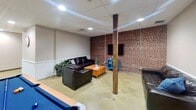 This 1220 square foot single-floor space in York, PA, is a multifunctional recreational area that combines comfort with entertainment. The room features a striking contrast of exposed red brick walls against white painted surfaces, creating a modern yet rustic ambiance. Recessed lighting in the drop ceiling provides ample illumination throughout.

The space is divided into distinct zones for various activities. A billiards area is centered around a high-quality pool table with blue felt, inviting casual games. Comfortable black leather sofas and armchairs are strategically placed, offering relaxed seating for conversation or spectating. A wooden coffee table adds a warm touch to the lounge area.

Industrial elements are evident in the exposed wooden support beam and concrete flooring, which has been polished to a smooth finish. Large glass doors and windows allow natural light to flood in, connecting the interior to the outside world. The room also includes additional seating options with cushioned chairs along the walls.

Practical amenities are not overlooked, with visible power outlets and what appears to be a kitchenette or bar area. The space is designed for versatility, suitable for social gatherings, recreational activities, or as a communal area in a larger facility. Its open layout and durable finishes make it adaptable for various uses while maintaining a welcoming atmosphere.