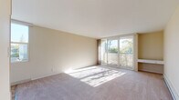 This single-floor residence is thoughtfully designed to maximize natural light and openness throughout. The spacious living area features soft wall-to-wall carpeting and warm, neutral-toned walls that create an inviting ambiance. A large sliding glass door opens directly to a private balcony, flooding the room with sunlight and offering pleasant views of the surrounding tree canopy. A built-in bench beneath the window further enhances the living space's functionality.

The bedroom is generously proportioned and features a wide bank of windows spanning nearly the full width of one wall, framing beautiful views of mature trees with seasonal foliage. Carpet flooring and neutral walls contribute to a calm, restful atmosphere.

An additional room, which could serve as a kitchen or dining area, features tile flooring and includes built-in cabinetry with overhead shelving and a countertop workspace, along with a ceiling fan for added comfort. This versatile space connects seamlessly to the rest of the unit.

The bathroom is clean and functional, finished with tile flooring, a white vanity with a large mirror, and a grab bar for accessibility. Throughout the residence, the consistent use of light, neutral tones and abundant glazing creates a cohesive and welcoming environment.