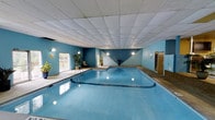 This indoor pool facility is a self-contained aquatic space designed for year-round use. The centerpiece is a generously sized rectangular swimming pool with clear blue water, illuminated by underwater lighting that enhances its inviting appearance. The pool is framed by a smooth concrete deck that provides ample surrounding space for movement and relaxation.

The room features bold blue walls accented with geometric design details, giving the space a distinctive and energetic character. A dropped tile ceiling adds a clean, structured aesthetic overhead, while wall-mounted lighting fixtures ensure the area is well lit throughout.

Lush potted plants are positioned around the perimeter, introducing a natural element that softens the room's hard surfaces and adds a touch of warmth to the environment. A seating area near the poolside offers a comfortable spot for rest and observation.

Large sliding glass doors along one wall open to an outdoor area, allowing natural light to filter in and providing easy access to the exterior. On the opposite side of the room, a door leads to additional amenities, suggesting the facility is part of a larger complex. Overall, the space strikes a balance between functionality and comfort, making it well-suited for recreational swimming and leisure activities.