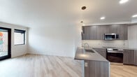 This contemporary single-floor unit showcases a thoughtfully designed open-concept layout that seamlessly connects the kitchen and living area. The kitchen is a standout feature, equipped with dark wood-grain cabinetry, a subway tile backsplash, stainless steel appliances including a refrigerator, range, and microwave, and a generous island with a dark quartz countertop and integrated sink. Sleek pendant lighting hangs above the island, complementing the recessed ceiling lights throughout. Light-toned hardwood-style flooring runs through the main living area, adding warmth and continuity to the space. The bedrooms are carpeted in a neutral tone, offering a comfortable contrast to the hard floors elsewhere. Large black-framed windows in the bedrooms allow natural light to fill the rooms while providing views of the surrounding modern architecture. The bathroom features a clean, minimalist design with light wood cabinetry, a white countertop, an undermount sink, and a large wall mirror. Crisp white walls throughout the unit enhance the bright, airy atmosphere. The overall aesthetic is modern and neutral, creating a versatile canvas well-suited to a variety of interior styles.