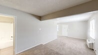 This single-floor unit offers a functional and efficiently laid out living space comprising a living room, kitchen, bathroom, and closet. The living room is generously proportioned relative to the overall footprint, featuring wall-to-wall beige carpeting, neutral warm-toned walls, and a textured popcorn ceiling. A wall-mounted HVAC unit and a window with blinds provide climate control and natural light, contributing to a bright and comfortable atmosphere. The front entry door opens directly into this open living area, which flows seamlessly throughout the unit. The kitchen is designed in a galley style, featuring crisp white shaker-style cabinetry with brushed nickel hardware, light stone-look countertops, and light wood-look tile flooring. Black and stainless steel appliances, including a refrigerator, range, and dishwasher, are integrated into the layout, offering a practical and clean aesthetic. The bathroom is finished with light wood-look tile flooring, a full-size tub and shower combination with a white surround, and clean white fixtures. A dedicated closet provides additional storage. The overall design maintains a consistent neutral palette of whites and warm grays, creating a clean, move-in-ready environment throughout.