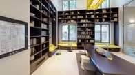 Located at 8560 Second Ave in Silver Spring, Maryland, this thoughtfully designed amenity space combines functionality with contemporary style across a single floor. The grand lobby entrance sets the tone immediately, featuring soaring floor-to-ceiling glass walls that flood the space with natural light, light-toned tile flooring, and an eye-catching sculptural ceiling installation that adds an artistic focal point overhead. A mounted display screen and clean-lined wall sconces contribute to the modern, welcoming atmosphere.

Beyond the lobby, a sophisticated library and lounge area offers an inviting retreat. Floor-to-ceiling dark wood bookshelves line the walls, filled with books and curated décor. Built-in window seating with bold yellow cushions provides comfortable spots to read or relax, with views of mature trees outside. A long communal work table with contemporary seating anchors the center of the room.

Adjacent to the lounge, a glass-walled meeting room with geometric-patterned frosted panels offers semi-private gathering space. Inside, yellow accent chairs surround a sleek conference table beneath stylish pendant lighting. Curated framed artwork adorns the walls throughout, reinforcing the space's polished, design-forward character. The consistent use of yellow accents, neutral tones, and natural materials creates a cohesive and energizing environment throughout.