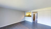 This single-floor residence offers a clean, functional layout designed for comfortable everyday living. The main living area is open and airy, featuring large-format dark tile flooring that adds a modern, polished aesthetic to the space. Neutral gray walls and white trim create a bright, neutral backdrop that lends itself well to a variety of interior styles.

The kitchen is positioned at the heart of the home, separated from the living area by a half-wall breakfast bar with a dark countertop. White cabinetry lines the kitchen walls, providing ample storage, while stainless steel appliances — including a refrigerator and microwave — give the space a contemporary feel. Track lighting illuminates the kitchen, keeping the work area bright and well-lit.

A hallway visible beyond the kitchen suggests additional rooms, likely including a bedroom and bathroom, continuing the home's consistent use of tile flooring and light tones. The overall ambiance of the space is clean and move-in ready, with a straightforward, practical design that maximizes the available footprint. The open sightlines between the living and kitchen areas make the home feel connected and spacious, ideal for both everyday living and casual entertaining.