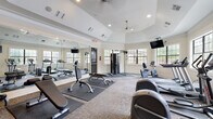 This fitness center is thoughtfully designed to support a comprehensive workout experience within a single-floor layout. The space features an elegant tray ceiling with recessed lighting and a ceiling fan, lending the room an open, airy feel. Large multi-pane windows line the walls, flooding the interior with natural light and offering views of the surrounding outdoor environment.

The cardio zone is well-appointed with multiple treadmills, elliptical trainers, and stationary bikes, all positioned to take advantage of the natural light and wall-mounted flat-screen televisions for entertainment during workouts. A dedicated strength training area includes a cable machine, adjustable weight benches, and rubber floor mats for safety and comfort.

The free weights section offers a full rack of dumbbells alongside additional benches, complemented by a large wall mirror that visually expands the space and allows users to monitor their form. The flooring throughout combines durable carpet tile with protective rubber mats under equipment zones.

The overall ambiance is clean, bright, and functional, with warm neutral wall tones and tasteful artwork adding character. A body weight scale and organized storage shelving round out the practical amenities, making this fitness center a complete and inviting space for users of all fitness levels.