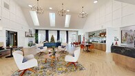 Located at 7800 Creekridge Rd in Charlotte, NC, this single-floor community clubhouse serves as the welcoming hub of a multi-building residential complex. The interior is defined by soaring vaulted ceilings fitted with large skylights that flood the open-plan space with natural light, complementing the light herringbone-patterned hardwood flooring throughout. A café-style bar area with warm wood cabinetry and subway tile backsplash offers a casual gathering spot, while nearby dining tables and chairs provide additional seating. A dedicated lounge zone features a curated arrangement of contemporary chairs and sofas, anchored by patterned area rugs. A banquette seating wall, adorned with bold, colorful artwork and flanked by round tables and accent chairs, adds a refined social area. Two bathrooms and a connecting hallway complete the interior layout. Externally, the property is part of a well-maintained multi-story residential complex clad in yellow siding and brick, featuring private balconies and ample surface parking. A fenced outdoor playground with a multi-structure play set sits adjacent to a wooded treeline, offering a natural backdrop and recreational space for residents.