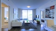 This single-floor residence showcases a clean, modern aesthetic centered around a well-appointed living area. The space features dark hardwood flooring that contrasts elegantly with the bright white walls and ceiling, creating a crisp, contemporary atmosphere. A large sliding glass door floods the room with natural light and opens to a private balcony, offering outdoor views and enhancing the sense of openness.

The living area is designed for both comfort and style, accommodating a white sectional sofa adorned with blue and white accent pillows, complemented by a rectangular wood-and-glass coffee table and a plush dark area rug. A wall-mounted flat-screen television sits above a low-profile media console, which features warm wood tones and decorative blue and green glass accents that add a pop of color. Vibrant abstract artwork lines one wall, contributing to the space's energetic yet relaxed ambiance.

A flush-mount ceiling light provides ambient illumination, while a partially visible adjacent room suggests additional living quarters. Overall, the space balances modern design with functional living, emphasized by its bright, airy quality and thoughtful décor choices.
