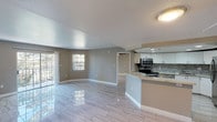 This single-floor residence offers an open-concept design that seamlessly integrates the living and kitchen areas, creating a bright and functional layout. The polished large-format tile flooring runs throughout the main living space, reflecting light and lending an airy, refined feel to the interior.

The kitchen is a standout feature, outfitted with a full suite of stainless steel appliances, including a range with over-the-range microwave, dishwasher, and a full-size refrigerator. Crisp white cabinetry extends to the ceiling and is complemented by a decorative tile mosaic backsplash that adds texture and visual interest. Light granite countertops provide ample prep and serving surface, while a breakfast bar peninsula creates a natural transition into the living area, ideal for casual dining or entertaining.

The living space is generously sized, featuring neutral warm-gray walls, clean white trim, and sliding glass doors that open to a private balcony, allowing natural light to fill the room. A secondary window provides additional brightness, and a doorway leads further into the residence. Recessed lighting in the kitchen and a flush ceiling fixture in the main living area ensure well-distributed illumination throughout. The overall ambiance is clean, modern, and move-in ready.