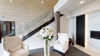 This refined lobby makes a strong first impression through its thoughtful blend of modern and classic design elements. The space is anchored by soaring ceilings that lend an airy, grand atmosphere, while large-format light stone tile cladding the walls adds warmth and texture throughout. A central staircase serves as the focal point of the lobby, featuring a sleek glass balustrade paired with a dark wood handrail and structural column, creating a refined contrast against the neutral stone backdrop. Pendant globe lights hang from above, casting a soft, ambient glow across the space. At the heart of the lobby, a welcoming seating area is arranged with a pair of cream upholstered wingback chairs finished with nailhead trim, positioned around a round table. A tall, elegant vase holding a lush arrangement of white floral blooms serves as a striking centerpiece, adding a fresh and sophisticated touch. Light-toned tile flooring grounds the space and complements the overall neutral palette, while a dark area rug beneath the seating area defines the conversation zone. The combination of modern glass detailing, rich wood accents, stone wall treatment, and curated furnishings creates a polished, welcoming environment that balances professionalism with understated elegance.