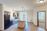 Located at 1004 Purple Iris Trail in Madison, Wisconsin, this single-floor residence offers a thoughtfully designed layout with one bedroom, one bathroom, and a dedicated closet. The open-concept living and kitchen area is a standout feature, showcasing light wood-style flooring, neutral walls, and abundant natural light streaming through vertical-blind-covered sliding glass doors and windows. The kitchen is well-equipped with rich, warm-toned cabinetry, black appliances including a refrigerator, range, microwave, and dishwasher, a double sink, and granite-style countertops. A central kitchen island with a matching countertop provides additional prep space and serves as a natural divider between the kitchen and living areas. Pendant lighting above the island adds a polished touch. The bedroom features plush carpeting and a bright window with blinds, offering a comfortable and quiet retreat. The closet provides practical storage adjacent to the bedroom. The bathroom includes a wide vanity with a large mirror, a well-lit fixture, tile flooring, and a clean, functional layout. Overall, the space presents a clean, modern aesthetic with a practical and efficient floor plan suited for comfortable everyday living.