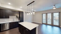 This thoughtfully designed one-bedroom, one-bathroom residence offers a generous and functional floor plan across a single level. The open-concept living and kitchen area serves as the heart of the home, anchored by a large center island with a crisp white countertop and a striking four-pendant light fixture overhead. The kitchen is outfitted with dark espresso shaker cabinetry, stainless steel appliances including a refrigerator, range, and built-in microwave, and a classic subway tile backsplash. Dark hardwood-style flooring flows throughout the main living areas, adding warmth and continuity. The spacious living room benefits from natural light through sliding glass doors that open to a private balcony, and is further enhanced by a ceiling fan. The bedroom features plush carpeting, a large window with exterior views, and access to a well-sized closet with built-in shelving. The bathroom is finished with marble-style tile flooring and wall surround, a deep soaking tub, dark vanity cabinetry, dual mirrors, and ample counter space. Neutral tones throughout the unit create a calm, move-in-ready ambiance suited to a variety of interior styles.