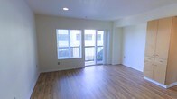 This single-floor residence offers a clean, contemporary layout with an open and airy feel throughout. The main living area is anchored by warm hardwood-style flooring that extends across the space, complementing the neutral, light-toned walls that create a welcoming and versatile backdrop. Recessed ceiling lighting provides consistent, even illumination, enhancing the brightness of the room. Natural light flows generously through a large window fitted with horizontal blinds, offering both privacy and light control. A sliding glass door leads directly to a private balcony, seamlessly connecting the interior living space to the outdoors. The balcony features metal railing and provides views of the surrounding exterior. The overall design is minimal and practical, with clean lines and a neutral palette that lends itself well to a variety of furnishing styles. The space presents a move-in ready condition with well-maintained finishes and thoughtful architectural details throughout, making it a functional and comfortable living environment.