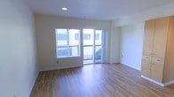 This single-floor residence offers a clean, contemporary layout with an open and airy feel throughout. The main living area is anchored by warm hardwood-style flooring that extends across the space, complementing the neutral, light-toned walls that create a welcoming and versatile backdrop. Recessed ceiling lighting provides consistent, even illumination, enhancing the brightness of the room. Natural light flows generously through a large window fitted with horizontal blinds, offering both privacy and light control. A sliding glass door leads directly to a private balcony, seamlessly connecting the interior living space to the outdoors. The balcony features metal railing and provides views of the surrounding exterior. The overall design is minimal and practical, with clean lines and a neutral palette that lends itself well to a variety of furnishing styles. The space presents a move-in ready condition with well-maintained finishes and thoughtful architectural details throughout, making it a functional and comfortable living environment.
