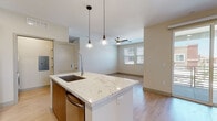 This well-appointed single-floor residence combines modern design with functional living across a thoughtfully laid-out floor plan. The open-concept kitchen is a standout feature, showcasing a large central island topped with striking granite countertops and an undermount sink with a sleek pull-down faucet. Rich wood-toned cabinetry extends to the ceiling, complemented by a classic white subway tile backsplash and a full suite of stainless steel appliances, including a refrigerator, range, and built-in microwave. Pendant lights hang above the island, adding warmth and style. Light hardwood flooring flows seamlessly from the kitchen into the adjoining living area, where a ceiling fan and window with blinds provide comfort and natural light. A sliding glass door opens to a private balcony, extending the living space outdoors. The bedrooms are carpeted and well-proportioned, each equipped with ceiling fans and windows that bring in natural light. The bathroom is finished with granite vanity counters, wood-framed mirrors, and a glass-enclosed walk-in shower with large-format tile. A dedicated laundry area houses a stacked washer and dryer unit alongside built-in shelving, offering ample storage. Throughout, neutral tones and clean lines create a cohesive and contemporary ambiance.