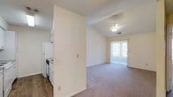 This single-floor residential space offers a practical and comfortable layout designed for everyday living. The kitchen is positioned adjacent to the main living area, separated by a partial wall that provides a sense of definition while maintaining an open feel. The kitchen is equipped with white appliances, including a refrigerator and stove, set against white cabinetry and contrasting dark wood-style flooring that adds a warm touch to the functional space.

The living area is carpeted and benefits from vaulted ceilings that lend a sense of openness and volume to the room. A ceiling fan is centrally mounted, providing both air circulation and a modest decorative element. Natural light fills the living space through a large sliding glass door fitted with vertical blinds, which also provides direct access to an outdoor area, enhancing the connection between interior and exterior.

The overall color palette is neutral and clean, with light-toned walls throughout that contribute to a bright and airy ambiance. The combination of vaulted ceilings, natural light, and an open flow between the kitchen and living area makes this a welcoming and functional single-floor residence.