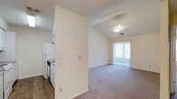 This single-floor residential space offers a practical and comfortable layout designed for everyday living. The kitchen is positioned adjacent to the main living area, separated by a partial wall that provides a sense of definition while maintaining an open feel. The kitchen is equipped with white appliances, including a refrigerator and stove, set against white cabinetry and contrasting dark wood-style flooring that adds a warm touch to the functional space.

The living area is carpeted and benefits from vaulted ceilings that lend a sense of openness and volume to the room. A ceiling fan is centrally mounted, providing both air circulation and a modest decorative element. Natural light fills the living space through a large sliding glass door fitted with vertical blinds, which also provides direct access to an outdoor area, enhancing the connection between interior and exterior.

The overall color palette is neutral and clean, with light-toned walls throughout that contribute to a bright and airy ambiance. The combination of vaulted ceilings, natural light, and an open flow between the kitchen and living area makes this a welcoming and functional single-floor residence.