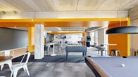 This generously sized amenity lounge offers a vibrant, open-plan layout defined by an industrial-modern aesthetic. Exposed concrete ceilings with visible ductwork and structural columns contrast boldly with striking yellow-orange accent beams and walls, creating an energetic atmosphere throughout the space. Large floor-to-ceiling windows and oversized glass sliding doors line multiple walls, flooding the interior with natural light and providing views of the surrounding streetscape and greenery.

The space is thoughtfully divided into distinct activity zones. A dedicated games area features a designer billiards table, a foosball table, a ping pong table, and an arcade gaming cabinet — all positioned to allow comfortable play without crowding. A lounge zone offers plush sofas, swivel chairs, and grouped seating arrangements around low tables, ideal for casual gatherings. High-top communal tables with metal barstools provide additional flexible seating for social interaction.

An integrated kitchen or kitchenette area with counter seating and appliances supports entertaining needs, while a glass-walled room offers a quieter, semi-private space within the larger open floor plan. Pendant lighting, track lighting, and ceiling-mounted fixtures complement the natural light throughout. The overall ambiance is lively, modern, and welcoming — well suited for socializing, recreation, and community engagement.