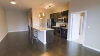 This single-floor space showcases a thoughtfully designed open-concept kitchen and living area that combines modern aesthetics with practical functionality. The kitchen is a standout feature, appointed with rich, dark espresso cabinetry that extends to the upper walls, creating a bold contrast against the light-toned countertops. Stainless steel appliances, including a gas range, microwave, and refrigerator, are seamlessly integrated into the design, lending a sleek, contemporary feel.

A central island serves as the focal point of the kitchen, offering additional counter space and casual bar seating with two wooden stools — ideal for dining or entertaining. A pendant light fixture hangs above the island, complementing the track lighting overhead that provides well-distributed illumination throughout the space.

Deep-toned hardwood flooring runs continuously across the open layout, anchoring the space with warmth and visual cohesion. The open sightlines between the kitchen and adjacent living areas enhance the sense of spaciousness. A doorway visible to the side suggests easy access to additional rooms, contributing to the overall functional flow of this well-appointed, modern interior.