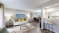 Located on Coronado Drive in Sarasota, Florida, this single-story residence presents a cohesive, coastal-inspired interior finished in soft neutral tones throughout. Wood-look luxury vinyl flooring runs continuously across the entire floor plan, providing a warm and unified foundation for each living space.

The open-plan living and dining area forms the heart of the home, where natural light filters in through a sliding glass door fitted with vertical blinds and drape panels. The living area offers generous wall space, highlighted by large-format coastal artwork, and flows seamlessly into a defined dining space illuminated by a pendant light fixture. A pass-through counter connects the dining area to the kitchen, which features a galley-style layout equipped with black appliances, including a range, refrigerator, and dishwasher, alongside white cabinetry and light countertops.

A secondary living or sitting area near the entry provides additional flexible space, accented by a wall-mounted television and white storage credenza. The bedrooms are quietly positioned away from the main living spaces, each featuring the same wood-look flooring, neutral walls, and a textured ceiling finish consistent throughout the home. The overall ambiance is bright, calm, and well-suited to the Florida lifestyle.